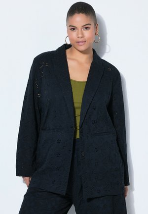 Mujer de cabello corto que lleva grandes pendientes de aro, blusa verde oliva y blazer de encaje floral negro con pantalones a juego sobre fondo blanco.