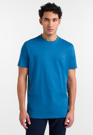 Młody mężczyzna o kręconych włosach, ubrany w gładki niebieski t-shirt z krótkim rękawem i ciemne spodnie, stojący na białym tle.