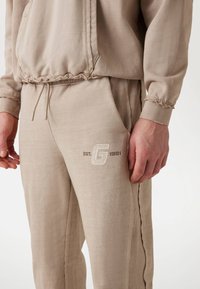 Beige szabadidőruha textúrázott felülettel. A felső részen elülső zseb és ráncolt alj található. A nadrágon hímzett logó és zsinóros derékpánt van.