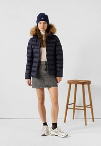 Giacca imbottita blu navy con cappuccio foderato di pelliccia, abbinata a un dolcevita crema, gonna in denim grigia e sneakers argento. Sgabello di legno nelle vicinanze.