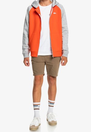 Young person wearing an orange and gray zip-up hoodie, white t-shirt, khaki shorts, striped socks, and white sneakers, standing against white background.