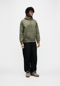 Φούτερ με κουκούλα σε ελιά πράσινο χρώμα με τσέπη τύπου καγκουρό, ριμπ μανσέτες και κορδόνι στην κουκούλα. Συνδυάζεται με μαύρο παντελόνι cargo και μπεζ μποτάκια με υφή στην σόλα.