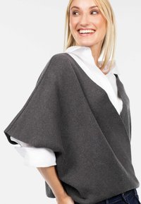 Pelēks oversized džemperis ar V veida kaklu, fakturētu adījumu un platām piedurknēm, kārtots virs baltas krekla ar apkakli. Ikdienas un mūsdienīgs izskats.