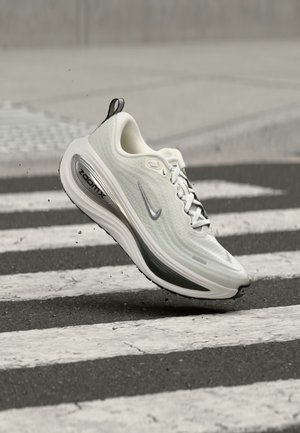 Chaussure de course Nike ZoomX blanche et grise en l’air au-dessus d’un passage piéton rayé noir et blanc sur une route asphaltée.