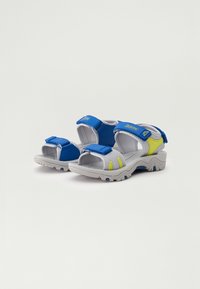 Zilās, pelēkās un dzeltenās sportiskās atvērtās sandales ar regulējamiem Velcro siksniņām un teksturētu gumijas zoli uz balta fona.