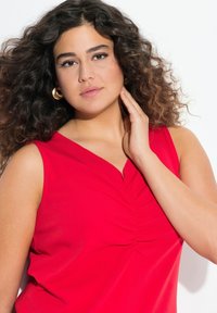 Rotes ärmelloses Top mit tiefem V-Ausschnitt, das in der Mitte mit Raffungen gestaltet ist. Glatte Textur, lebendige Farbe und minimalistisches Design.