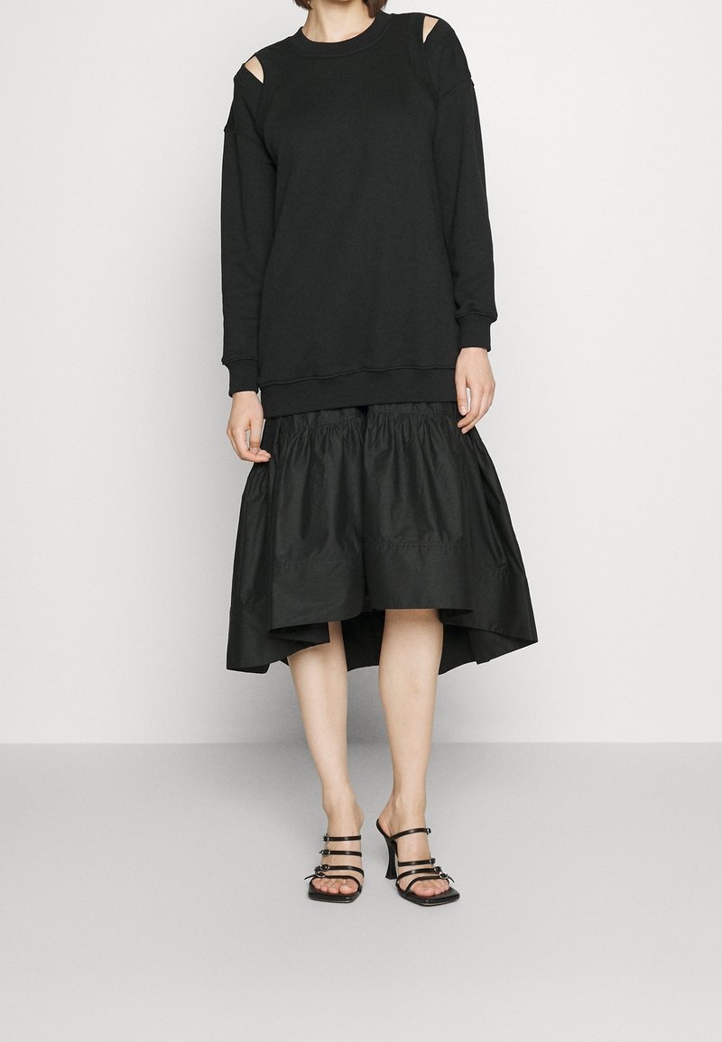 Femme portant un haut noir à manches longues avec découpes aux épaules, une jupe noire asymétrique et des sandales à talons noires à brides sur un sol gris.
