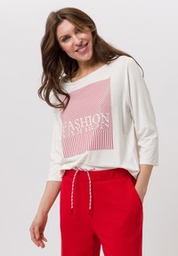 Haut à manches longues ample et blanc avec un motif graphique à rayures rouges, associé à un pantalon rouge à cordon de serrage. Le tissu semble doux et confortable.