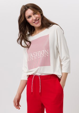 Haut à manches longues ample et blanc avec un motif graphique à rayures rouges, associé à un pantalon rouge à cordon de serrage. Le tissu semble doux et confortable.