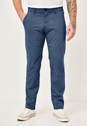 Man wearing navy blue slim-fit pants, white sneakers, and a white shirt, with tattooed right forearm visible.