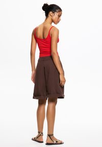 Red fitted tank top, brown knee-length shorts with rolled cuffs, and flat black sandals with a patterned strap. Simple and casual design.