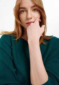 Teal ribbeltrui met wijde halslijn. Model heeft middel-lang haar en steunt met de hand op de kin, waarbij ze een gladde huid en natuurlijke make-up toont.