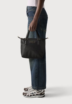 Borsa a mano nera in nylon con dettagli in pelle, logo dorato "REPLAY", due manici corti e chiusura con zip. Abbinata a jeans blu e sneakers.