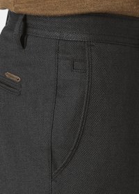 Black textured pants featuring a diagonal pocket with subtle stitching and a branded metal accent. The waistband appears elastic for comfort.
