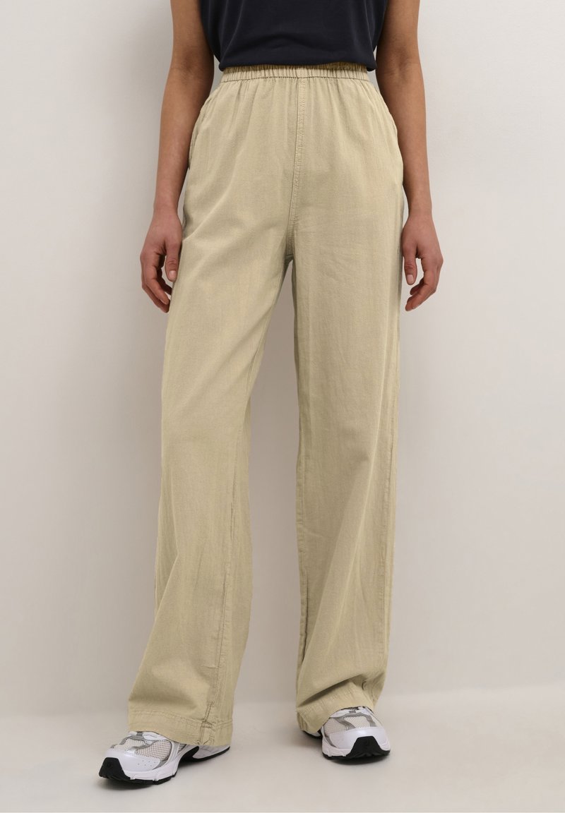 Ljusbeiga vida byxor med elastisk midja, tillverkade av mjukt tyg, i kombination med grå och vita sneakers. Slät textur, avslappnad design.