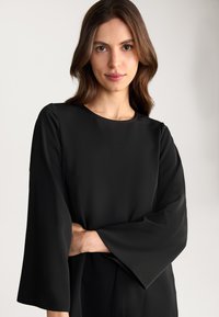 Zwarte blouse met lange mouwen, een ronde halslijn en brede uitlopende mouwen; gladde textuur en minimalistisch ontwerp.