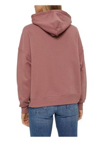 Femme portant un sweat à capuche ample mauve et un jean bleu, vue de dos avec les mains détendues le long du corps.