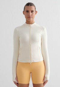Hvit zip-up treningsjakke med lange ermer og to sidelommer, kombinert med lyseoransje shorts, glatt tekstur, tettsittende design.