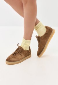 Legs wearing yellow socks and brown suede sneakers with gum soles, positioned against a plain light background.