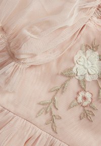 Helles rosa Gewebe mit transparenter Überlagerung, versehen mit floralem Stickerei in Creme- und Rottönen. Geraffter Rüschendetail am unteren Rand.