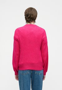 Cilvēks ar īsiem brūniem matiem, tērpies spoži rozā ribota džempera un zilu džinsu, stāv pretējā virzienā uz vienkrāsaina balta fona.