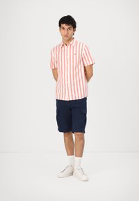 Kortermet skjorte med knapper i rosa og hvite vertikale striper, sammen med marineblå cargo-shorts, hvite sneakers og ankelsokker.