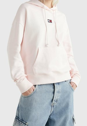 Sweatshirt à capuche rose clair avec une poche avant, des poignets côtelés et un petit logo noir et rouge sur la poitrine, fabriqué dans un matériau en coton doux.