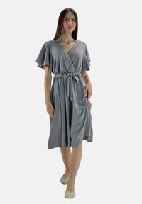 Robe grise à manches courtes et flottantes, nouée à la taille, avec une texture lisse et une longueur au genou. Portée avec des ballerines de couleur claire.