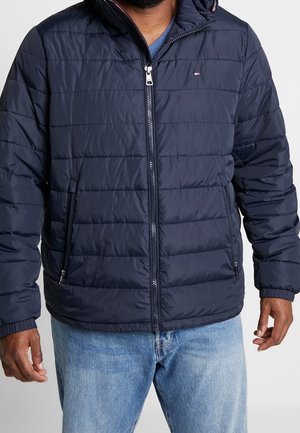 Homme portant une veste matelassée bleu marine avec une fermeture éclair à l'avant et des poches latérales zippées, associée à un jean bleu clair.