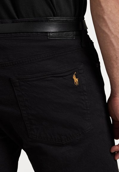 Calças de ganga pretas em algodão com um acabamento texturizado. Apresentam um bolso traseiro visível com um logótipo bordado amarelo em contraste. Costuras bem definidas.