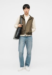 Młody mężczyzna w brązowej kamizelce zapinanej na zamek, na beżowej koszuli, niebieskich dżinsach i białych trampkach, niosący dużą czarną torbę na ramię.