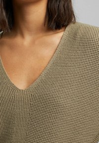 Gros plan sur une personne portant un pull à col en V vert olive texturé avec un motif en maille gaufrée, les cheveux courts et foncés visibles.