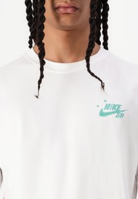 Weißes Langarm-T-Shirt aus Baumwolle mit einem türkisfarbenen Nike SB-Logo auf der linken Brust und einem Rundhalsausschnitt.