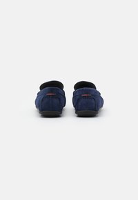 Mocassins en daim bleu marine avec un dos arrondi, présentant un accent de logo rouge et un détail de couture minimal. Semelle en caoutchouc noire à profil bas.
