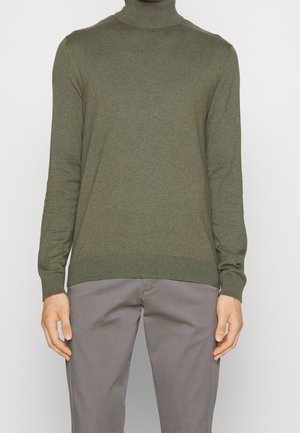 Homme portant un pull à col roulé vert olive et un pantalon gris, debout devant un fond clair uni.