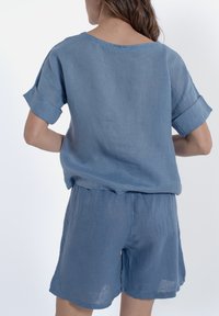 Top in lino blu con maniche corte arrotolate, abbinato a pantaloni corti coordinati. Vestibilità ampia, texture leggera e design semplice.