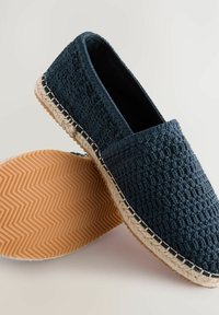 Espadrilles tejidas de color azul oscuro con una apertura elástica, suela de yute y un patrón de espiga en la suela de goma. Diseño plano y cerrado.