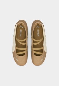 Ζευγάρι μπεζ μπαλαρίνες με λουράκια που διασταυρώνονται και λευκά λογότυπα Puma στο μπροστινό μέρος και στην εσωτερική σόλα, εμφανιζόμενες από πάνω σε λευκό φόντο.