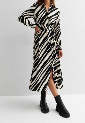 Kobieta ubrana w czarno-białą sukienkę kopertową w paski zebry z długim rękawem i guzikami oraz czarne, masywne botki na kostkę.