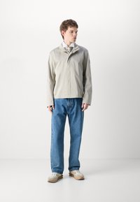 Lys grå vanntett jakke med høy krage, glidelås foran og lange ermer, sammen med løse blå denimjeans og nøytrale joggesko.