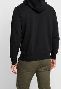 Felpa nera con una superficie liscia, cappuccio regolabile e polsini a costine. Indossata con pantaloni verde oliva, mostrando la vista posteriore.