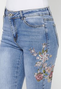 Light blue denim jeans with floral embroidery on the right thigh, featuring pink and green threads, five-pocket design, and light washing.