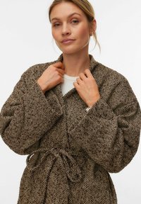 Brūna kažoka ar zivju kārtiņas rakstu, ar jostu, plašām piedurknēm un šaļveida apkakli, izgatavota no teksturētas auduma. Valkāta virs gaiša topa.