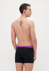 Boxer-briefs noirs avec une large ceinture violette marquée "BJÖRN BORG". Texture lisse, longueur mi-cuisse, avec un tatouage visible dans le dos.