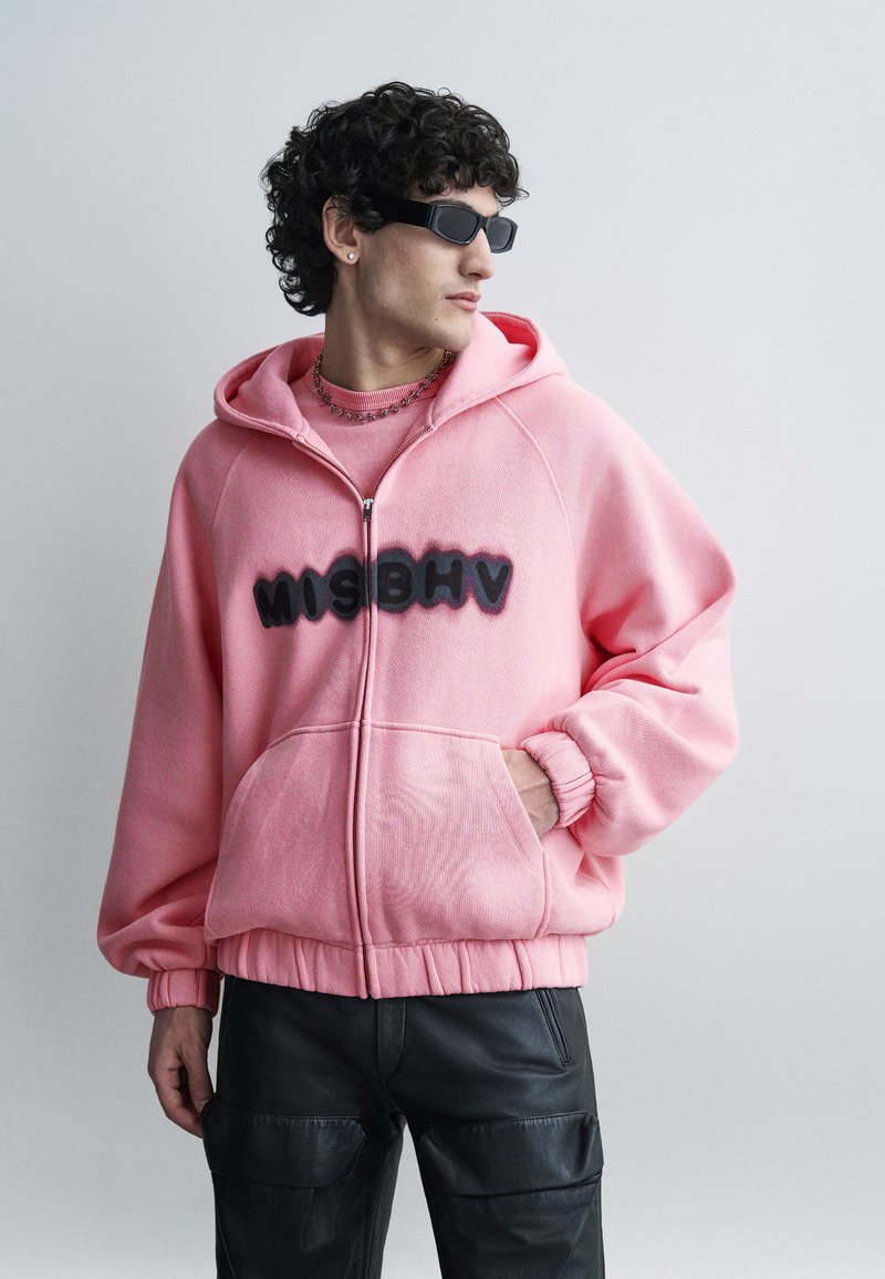 Giovane uomo con capelli ricci che indossa occhiali da sole neri, una felpa con cappuccio rosa con la scritta "MISBHV" e pantaloni cargo neri, che guarda di lato.