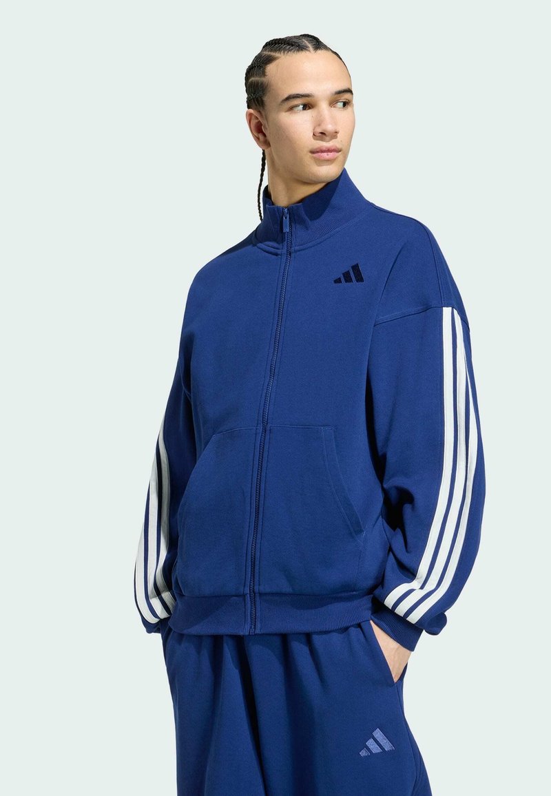 Jeune homme aux cheveux tressés portant un survêtement Adidas bleu à bandes blanches, debout les mains dans les poches, regardant sur le côté.