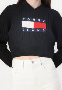 Czarny, żeberkowany, skrócony sweter z dekoltem w serek, z wielokolorową naszywką logo "TOMMY JEANS" w granatowym, czerwonym i białym kolorze z przodu.