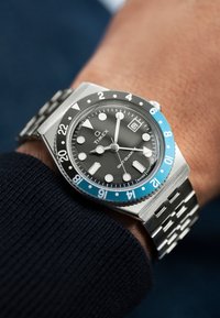 Montre en acier inoxydable avec un cadran noir, une lunette bleue et un bracelet en maillons argentés. Elle possède des marqueurs luminescents, un affichage de la date et un design audacieux.