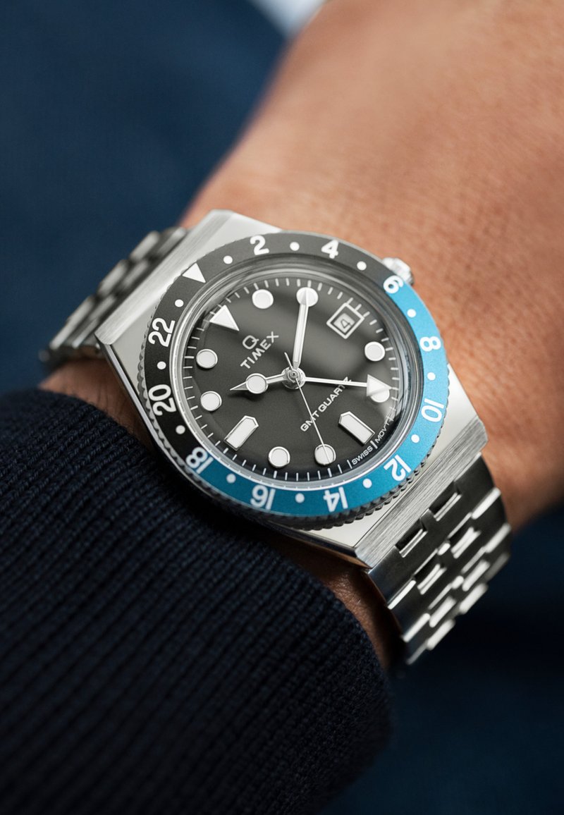 Montre en acier inoxydable avec un cadran noir, une lunette bleue et un bracelet en maillons argentés. Elle possède des marqueurs luminescents, un affichage de la date et un design audacieux.
