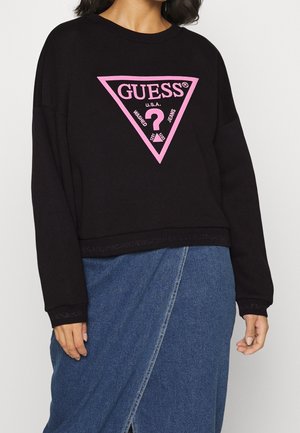 Personne portant un sweat-shirt noir Guess avec un logo triangle rose et une jupe portefeuille en denim bleu, debout devant un fond uni.